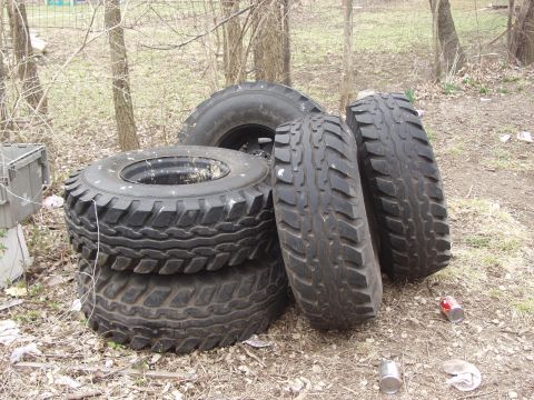 1979 Ford Bronco - A stack of four large, knobby off-road tires sits on the ground amidst trees and dry leaves.