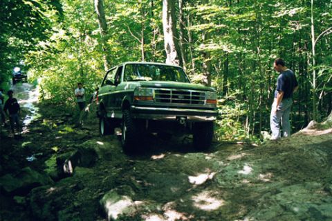 1995 Ford Bronco - A green 1995 Ford Bronco navigates a rocky, wooded trail with several people present and another vehicle in the background.
