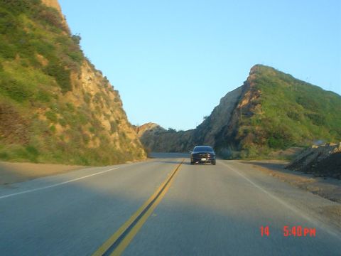 1993 Ford Taurus - A dark sedan drives down a two-lane road flanked by hills under a clear blue sky. A timestamp is visible in the lower right corner.