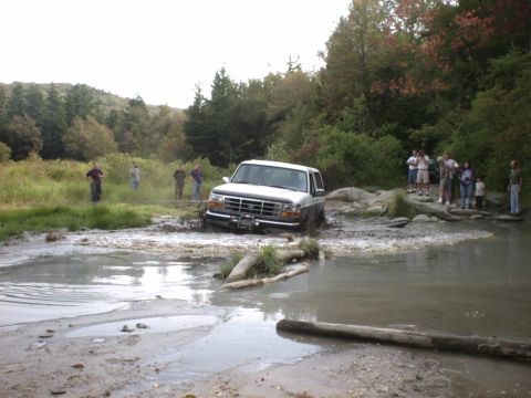 1995 Ford Bronco - A white Ford Bronco navigates a muddy off-road obstacle with onlookers present.