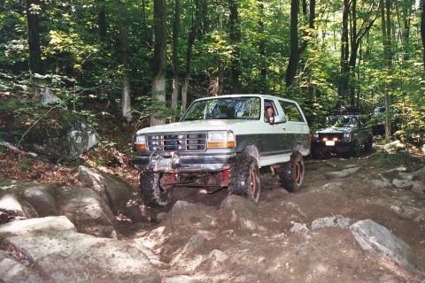 1995 Ford Bronco - Two SUVs, including a 1995 Ford Bronco, tackle a challenging rocky path in a wooded area.