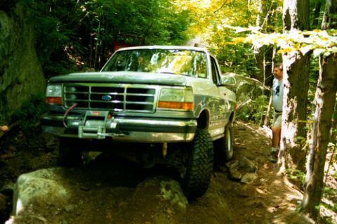 1995 Ford Bronco - A man watches as a Ford pickup truck navigates a challenging, rocky path through a dense forest.