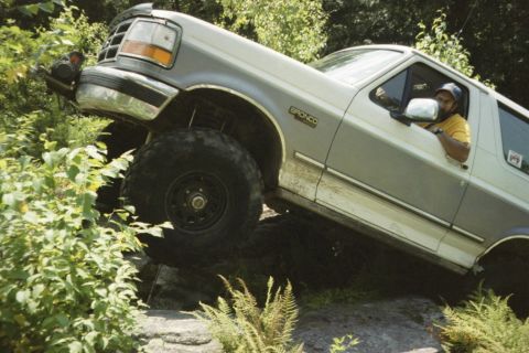 1995 Ford Bronco - A two-tone Ford Bronco is shown tackling a challenging off-road trail with large tires and a lifted suspension.