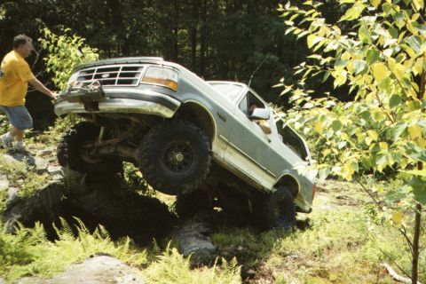 1995 Ford Bronco - A man in a yellow shirt pushes a gray Ford Bronco as it climbs over rocks.