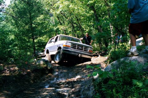 1995 Ford Bronco - A 1995 Ford Bronco tackles a challenging off-road course, with trees and rocks surrounding the muddy path.