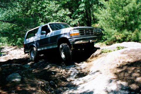 1995 Ford Bronco - This two-tone Ford Bronco is shown tackling a challenging off-road course with large rocks and dirt.