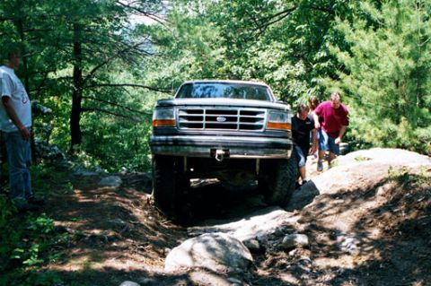 1995 Ford Bronco - A Ford Bronco drives off-road through a wooded area with several people watching.