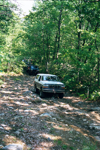 1995 Ford Bronco - A white Ford Bronco navigates a rough, unpaved path through a dense forest.
