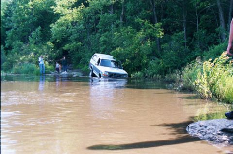 1995 Ford Bronco - An SUV is stuck in a body of water, with several people observing from the shore.