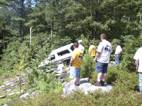 1995 Ford Bronco - A group of people observe a white SUV that has gone off the road and is lodged on a steep, wooded embankment.