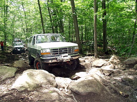 1995 Ford Bronco - A 1995 Ford Bronco with a winch tackles a challenging rocky trail in a wooded area, with another off-road vehicle following behind.