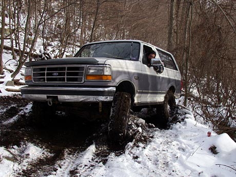 1995 Ford Bronco - A two-tone Ford Bronco navigates a snowy and muddy trail in a wooded area.