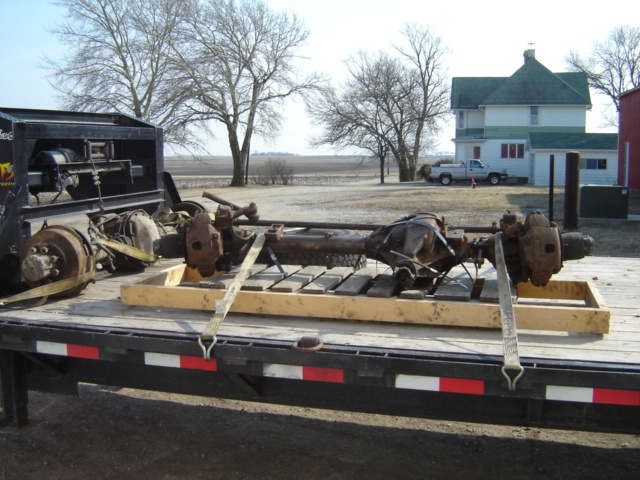 Front and rear axles loaded onto a flatbed trailer, with a house and trees in the background