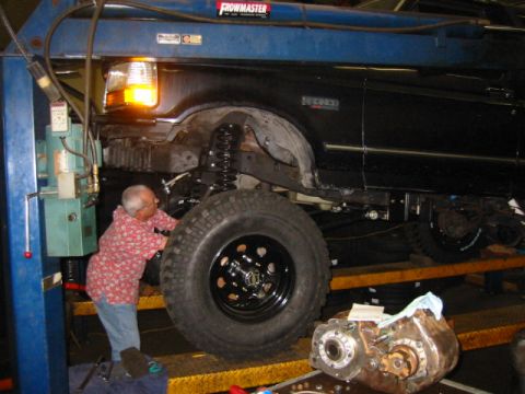 1992 Ford Bronco - A black Ford truck raised on a blue two-post shop lift with the front suspension drooped, showing an extended black coil spring, the expose…