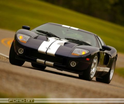 1985 Ford Bronco - A dark blue Ford GT with white racing stripes is captured in motion on a paved road.