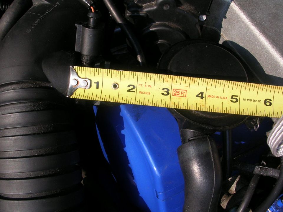 Yellow measuring tape across black hoses and a blue plastic component in an engine bay