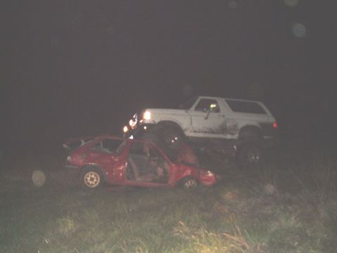 1989 Ford Bronco - White 1989 Ford Bronco perched atop a crushed red car in a nighttime scene.