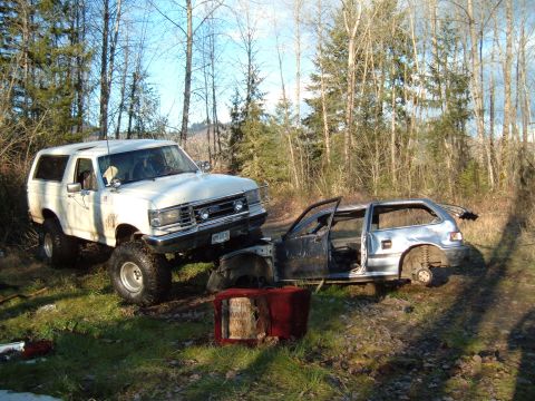 1989 Ford Bronco - A lifted white Ford Bronco is positioned precariously on top of a heavily damaged silver hatchback in a wooded area.