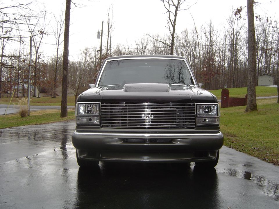 Front view of gray Bronco II with cowl hood and billet grille on wet driveway among bare trees