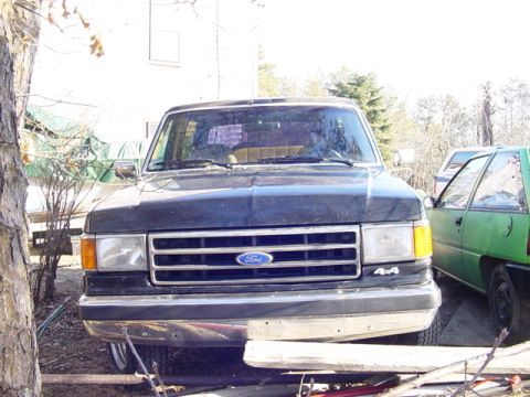 1989 Ford Bronco - A dark blue Ford Bronco from the late 1980s is seen from the front, parked next to a green car.