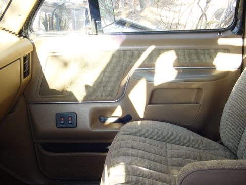 1989 Ford Bronco - The interior of a vehicle shows a tan door panel and a textured fabric seat, with sunlight casting shadows.