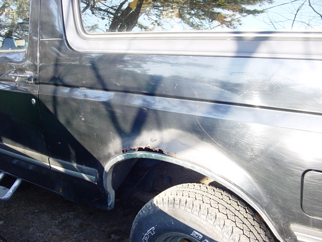 Side view of a dark-colored Ford Bronco showing rust around the rear wheel well and some body damage