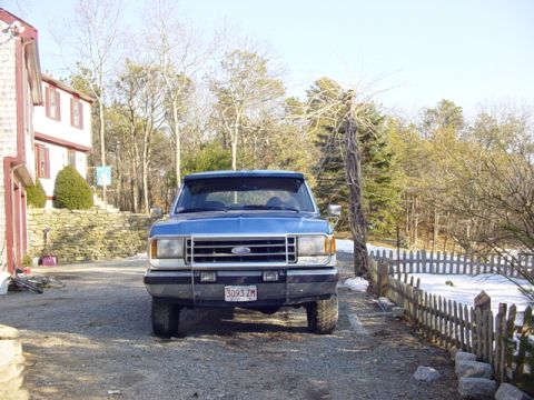 1989 Ford Bronco - A blue pickup truck is parked in a gravel driveway in front of a house with a wooden fence.