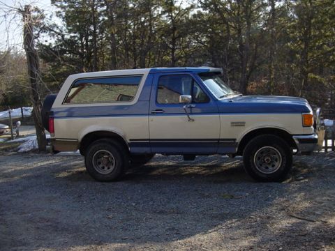 1989 Ford Bronco - This two-tone Ford Bronco is seen from the side, parked outdoors with trees and a fence visible.