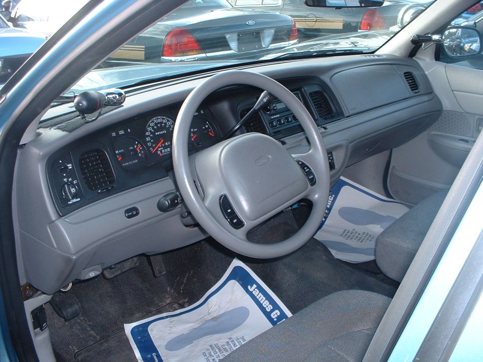 Interior view of a 1998 Ford Crown Victoria showing dashboard, steering wheel, and seats. Another Crown Victoria is visible through the windshield