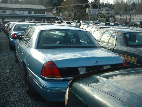 1999 Ford Crown Victoria - A row of sedans are parked on a gravel lot. The main car is light blue with a Ford emblem on the rear.