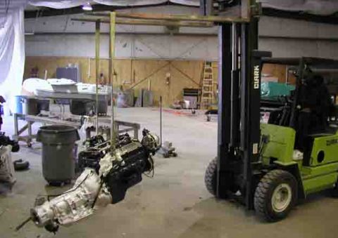 1988 Ford Bronco - A forklift lifts an engine and transmission assembly in a workshop setting, with a boat on a stand in the background.