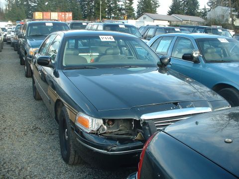 1999 Ford Crown Victoria - A dark sedan, likely a former police vehicle, is parked in a lot with visible damage to its front end.