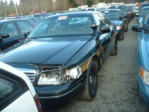 1999 Ford Crown Victoria - A dark sedan with a damaged front end is parked in a lot alongside other cars.