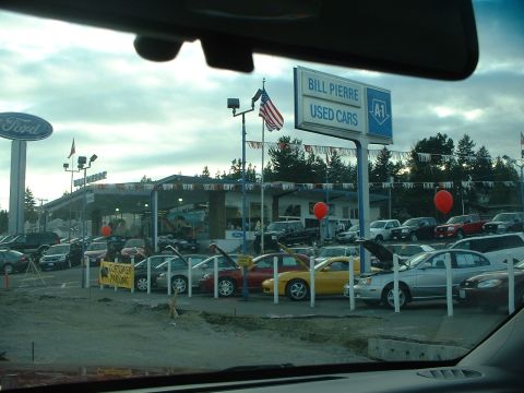 1999 Ford Crown Victoria - A view of a used car dealership lot with a prominent 'Bill Pierre Used Cars' sign and numerous vehicles parked under a cloudy sky.