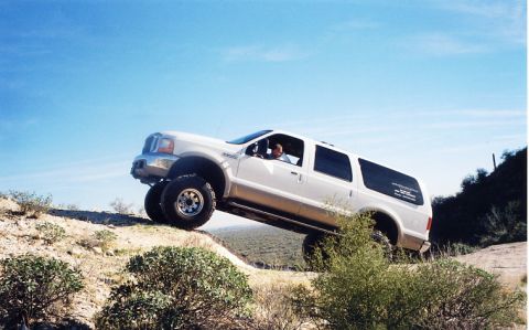 2001 Ford Excursion - A white Ford Excursion SUV navigates a steep dirt incline under a clear blue sky.