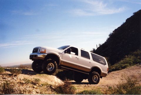2001 Ford Excursion - A white 2001 Ford Excursion SUV with a lifted suspension navigates a dirt incline under a clear blue sky.