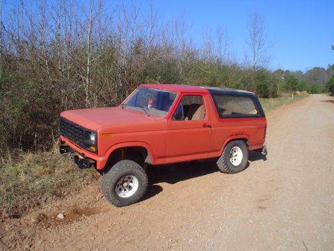 1981 Ford Bronco - This red Ford Bronco is parked on a dirt road, surrounded by trees under a clear blue sky.