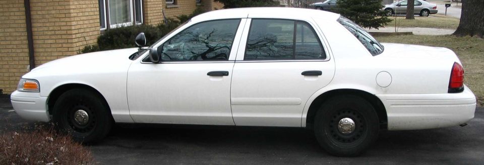 Side view of white Crown Victoria sedan with black steel wheels parked by a brick house