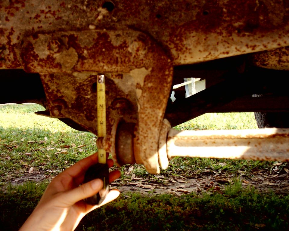 Rusted vehicle frame with yellow measuring tape held by hand, showing suspension component
