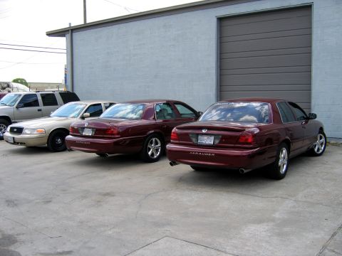 2000 Ford Crown Victoria - Several vehicles are parked in a row outside a commercial building with a large garage door.