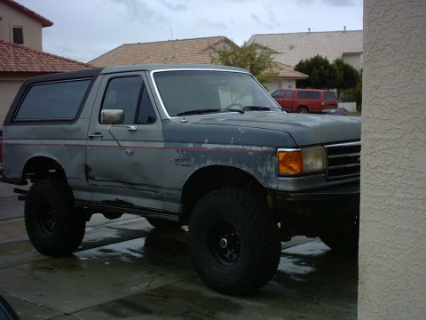 1990 Ford Bronco - A weathered gray Ford Bronco is parked in a driveway, with another red truck visible in the background.