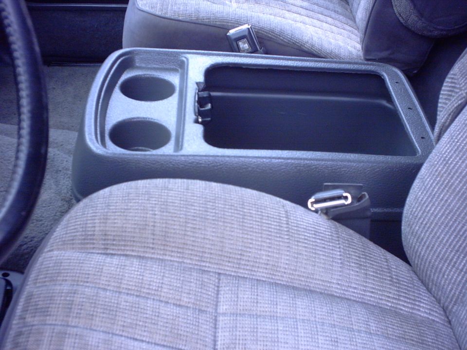 Interior shot of a vehicle's center console with cup holders and storage compartment, next to patterned seats and a seatbelt