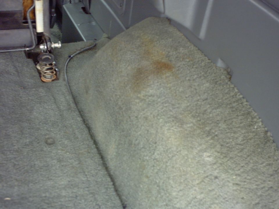 Carpeted rear interior of a vehicle, showing a seat bracket and some stains on the carpet