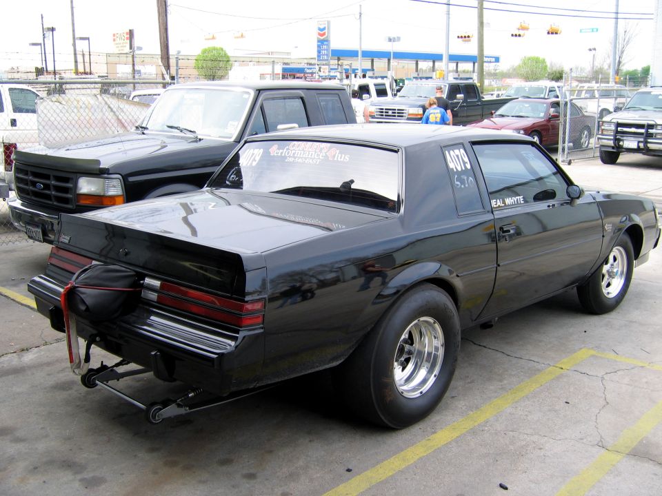 Black drag car with large rear tires and drag chute, parked next to a black Ford pickup truck