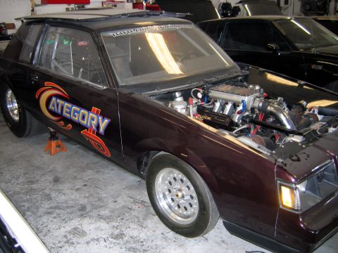 2000 Ford Crown Victoria - A modified drag car with custom graphics and an exposed engine sits in a garage next to another dark vehicle.