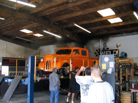2000 Ford Crown Victoria - An orange pickup truck is positioned on a dynamometer lift inside a workshop, with several people observing.