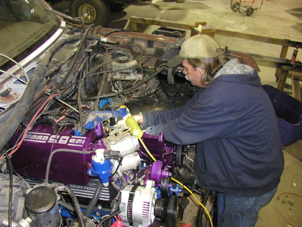 Man working on a 1988 Ford Bronco engine, which has purple valve covers reading '5.8 EFI'