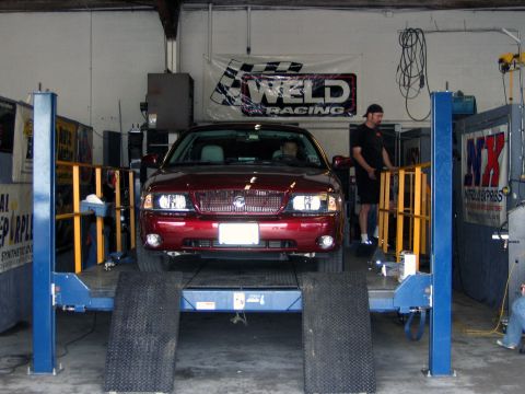 2000 Ford Crown Victoria - A red Mercury Grand Marquis is positioned on a lift inside a garage, with a Weld Racing banner visible overhead.