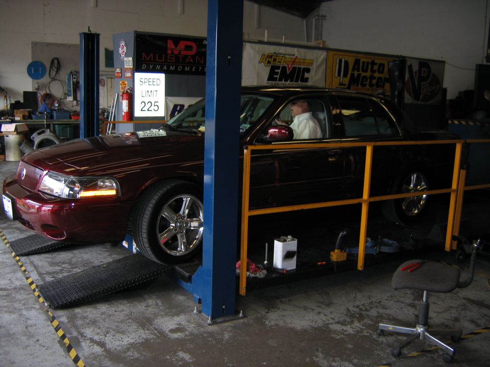 Maroon 2000 Ford Crown Victoria on a dynamometer lift in a garage. Speed limit sign reads 225