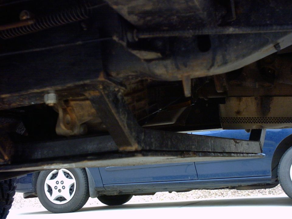 Undercarriage of a dark-colored vehicle with a blue car visible below, showing suspension components and a wheel with a star-shaped hubcap